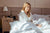 Woman in white pajamas holding Olverum Restful Sleep Pillow Mist, enjoying a peaceful morning in a cozy bedroom.